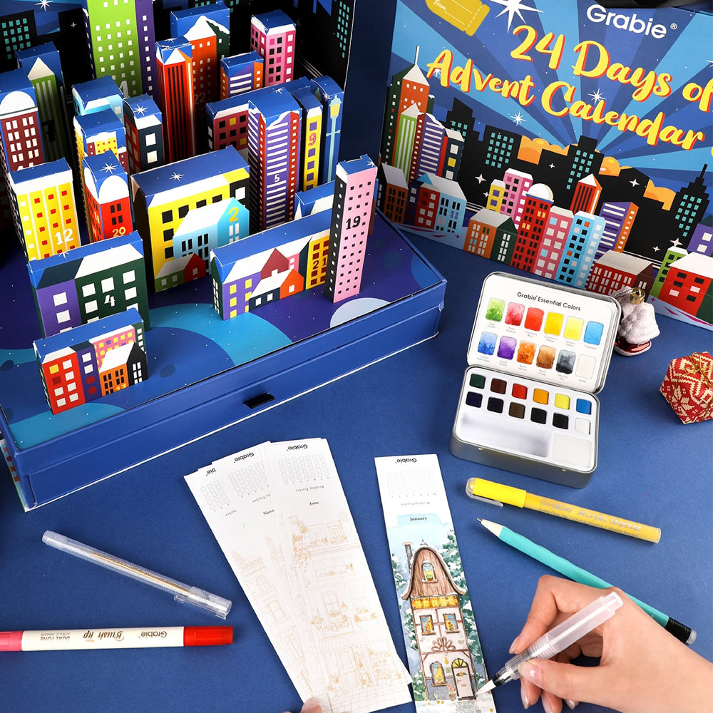 Advent calendar with miniature buildings, art supplies, and a hand holding a pen on a blue surface.
