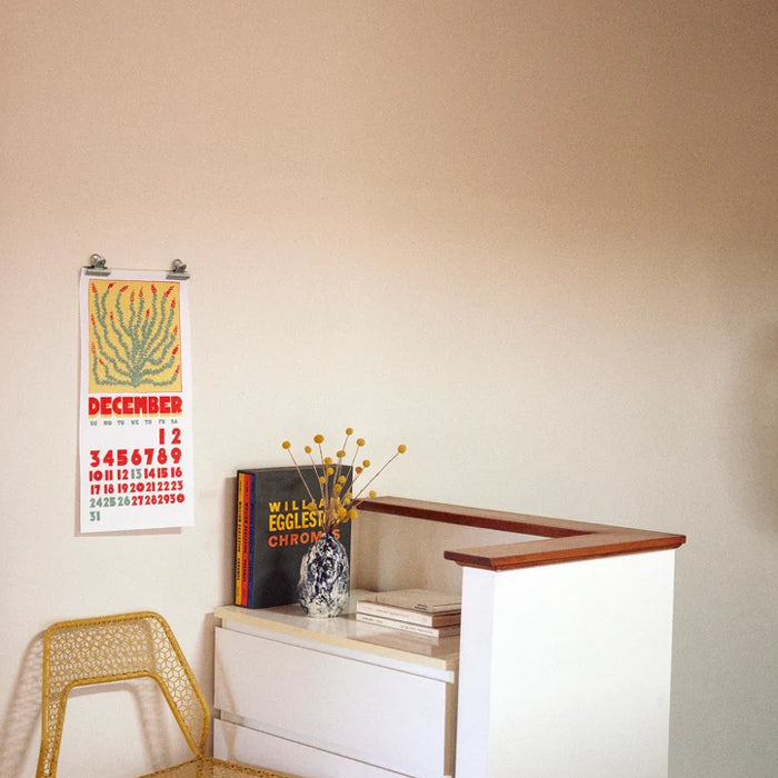 A vintage-style December calendar poster hangs on a beige wall above a white cabinet with a wooden top. The poster features a yellow-orange abstract tree design and red text.