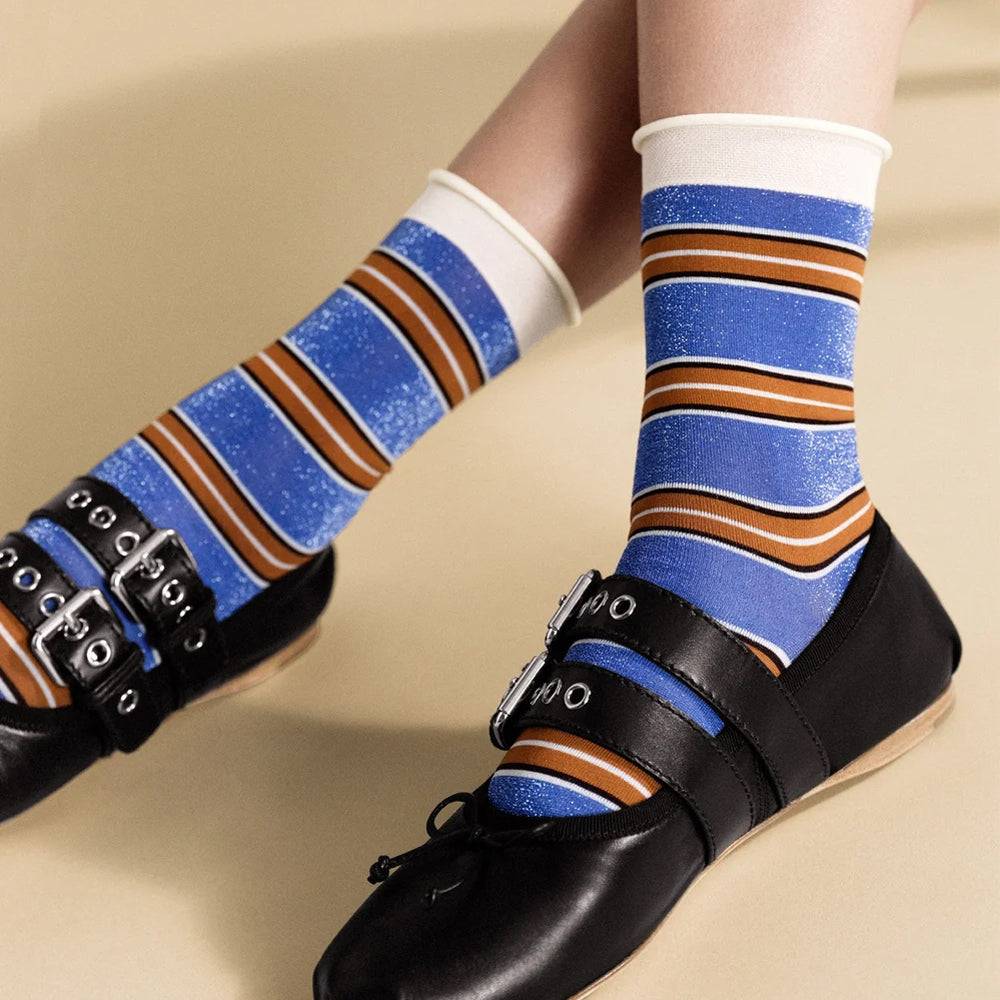 A close-up of a person's legs wearing blue, brown, and white striped socks with black double-strap Mary Jane shoes on a beige background.