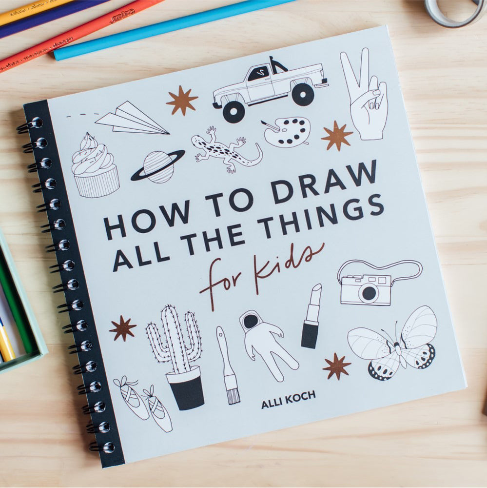 Book titled 'How to Draw All the Things for Kids' on a wooden surface with drawing illustrations.