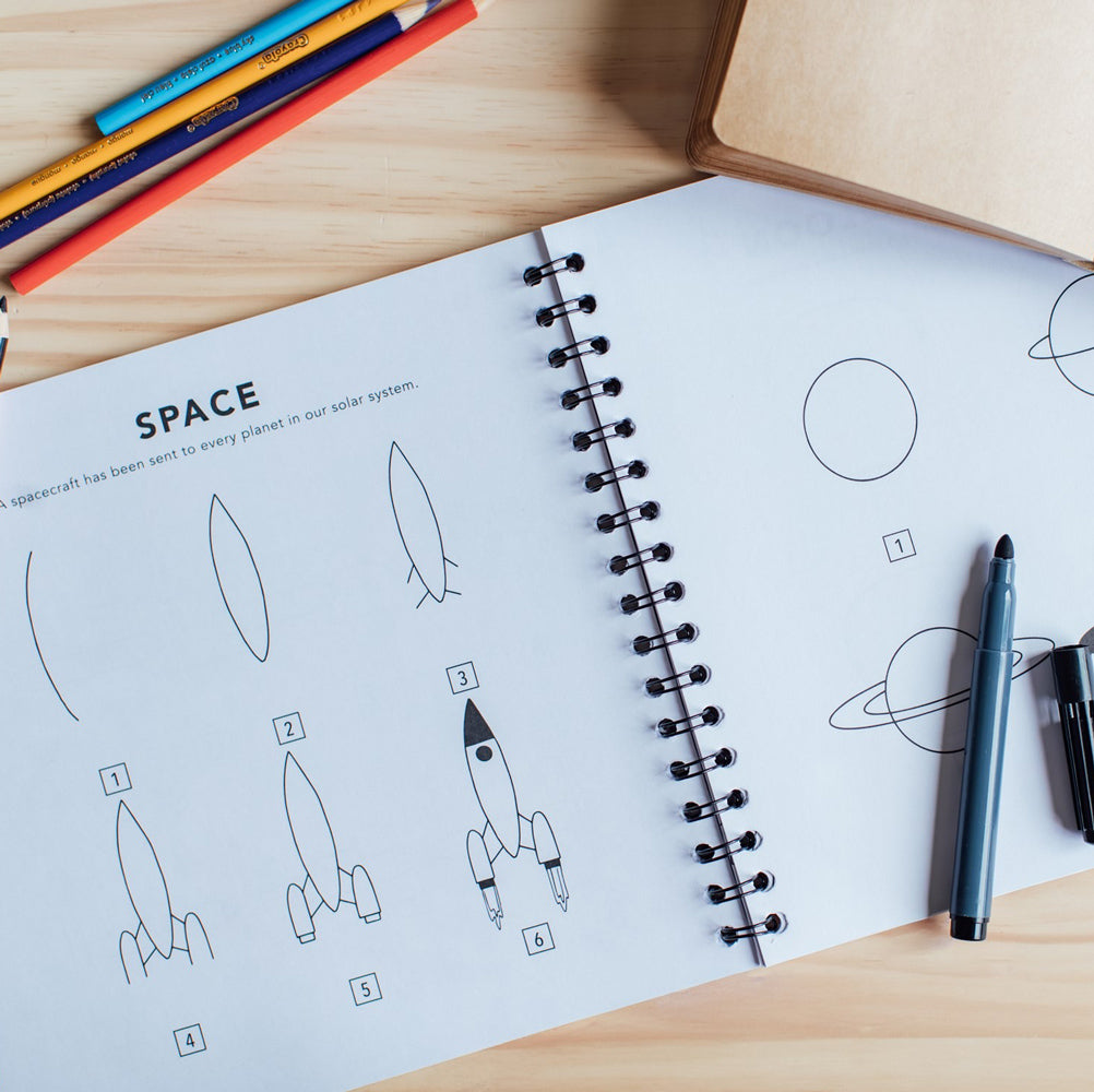 Interior page with space-themed drawing instructions and pencils on a wooden surface.