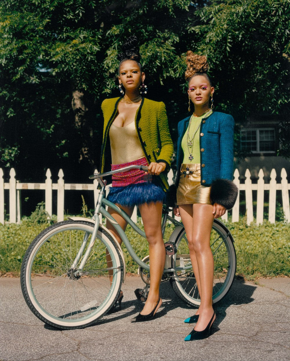 Two women standing next to a bicycle on a street with a white picket fence and greenery in the background.