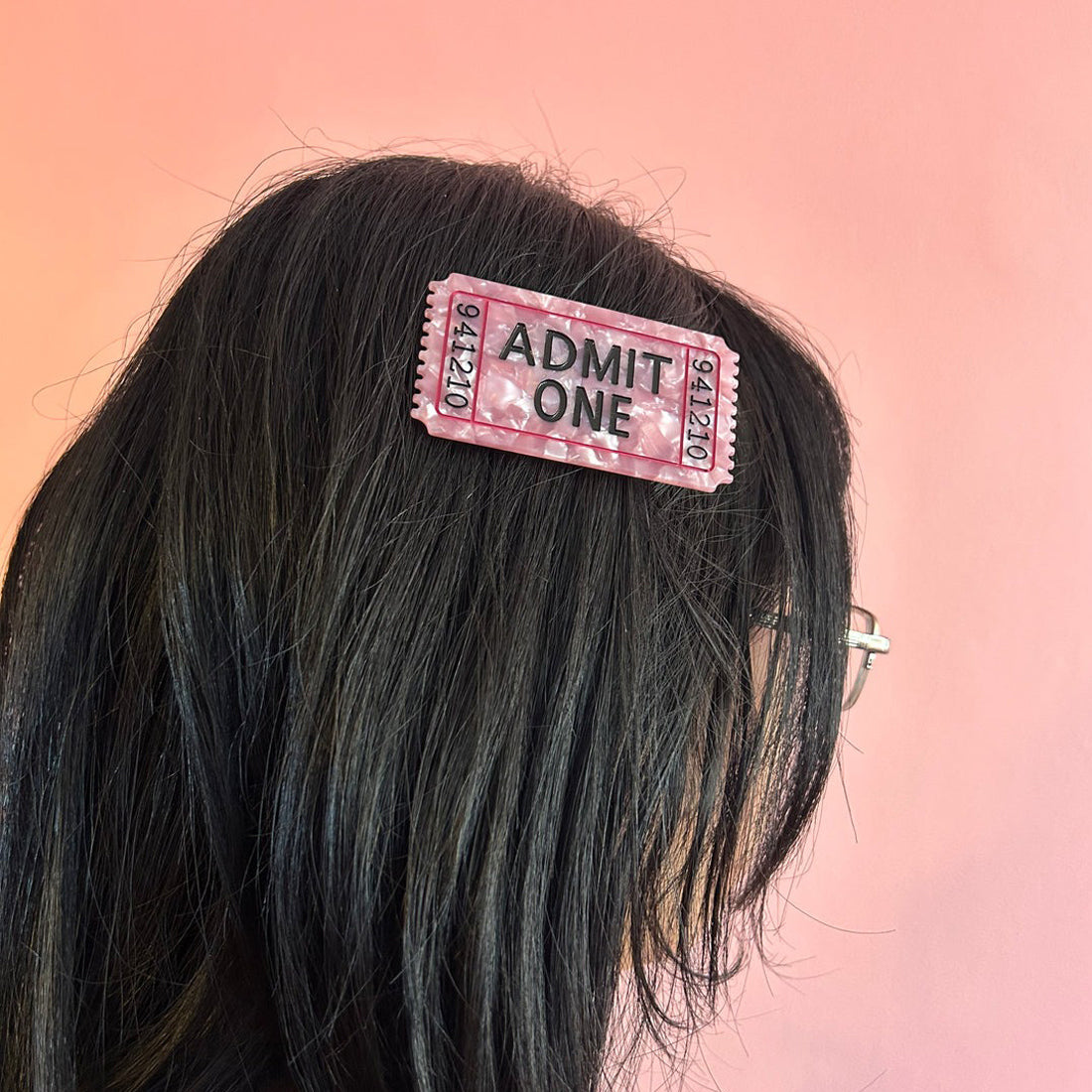Pink 'Admit One' ticket-shaped hair clip on black hair against a pink background