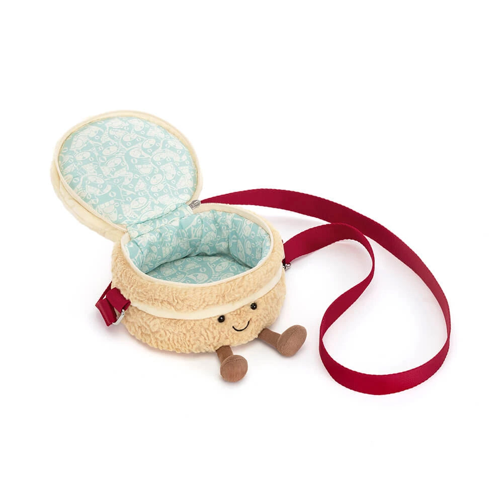 A plush bag shaped like a smiling cake, featuring a cream-colored textured exterior, a light blue patterned fabric lining, and a red shoulder strap, shown on a white background.