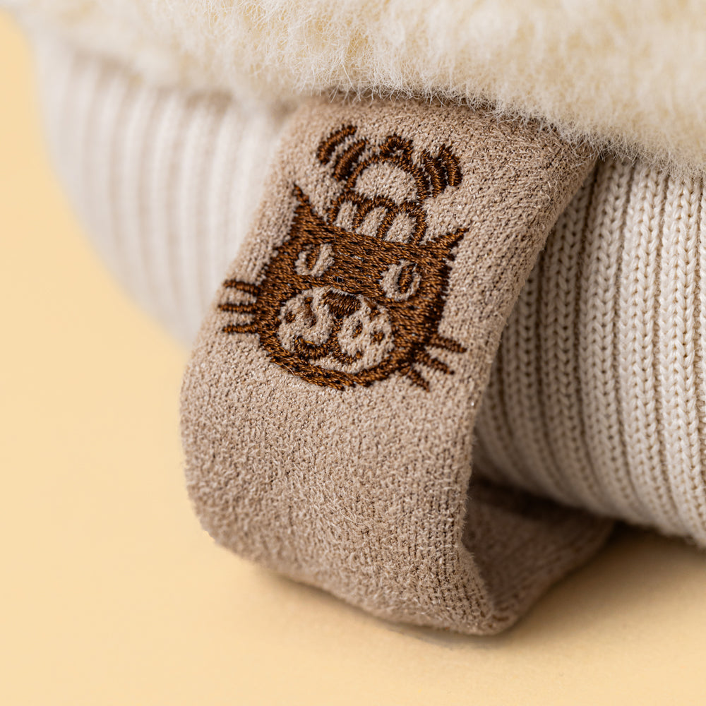 Close-up of a beige knit label with a brown embroidered cat face wearing a small hat, resting on a cream faux-fur surface against a warm beige background.