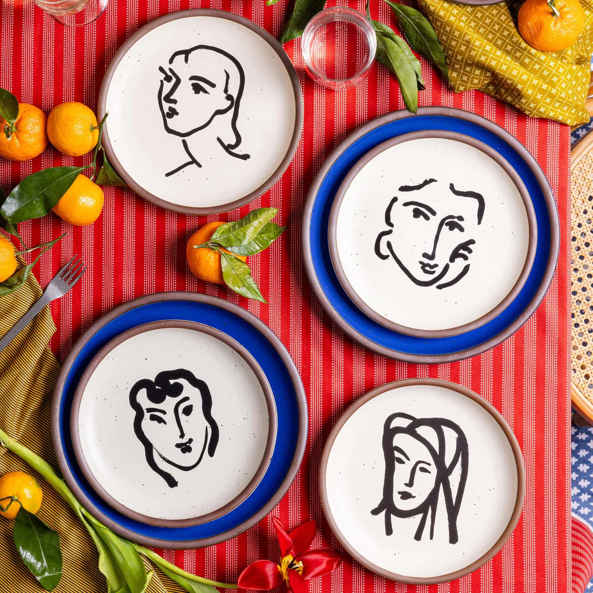Four ceramic plates with artistic face designs on a red tablecloth with fruits and leaves.