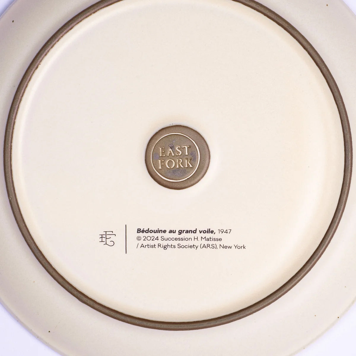 The underside of a ceramic plate or dish, showing a cream-colored glaze, a brown rim, and a central circular metallic emblem reading 'EAST FORK'. Below the emblem, printed text includes 'Bédouine au grand voile, 1947' and artist/copyright details.