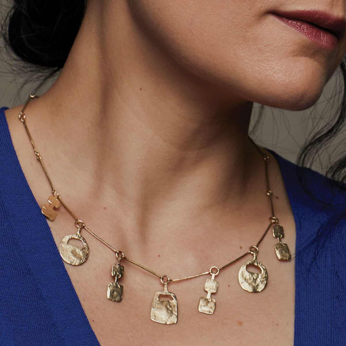 A gold necklace featuring a beaded chain with multiple dangling textured charms, including oval and rectangular pendants, displayed on a clean white background.