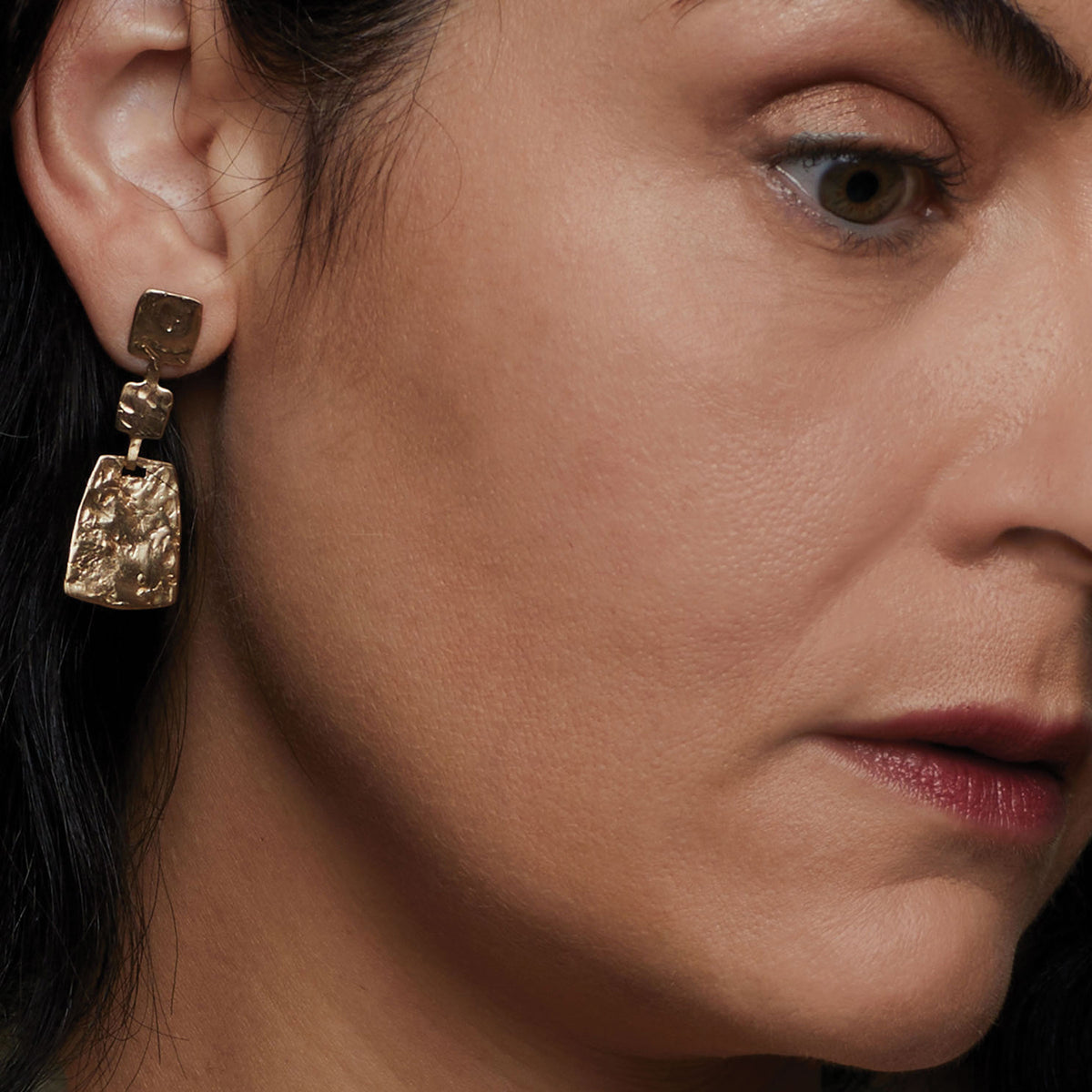 A close-up of a woman's ear wearing a textured gold drop earring with a square-shaped dangling pendant.