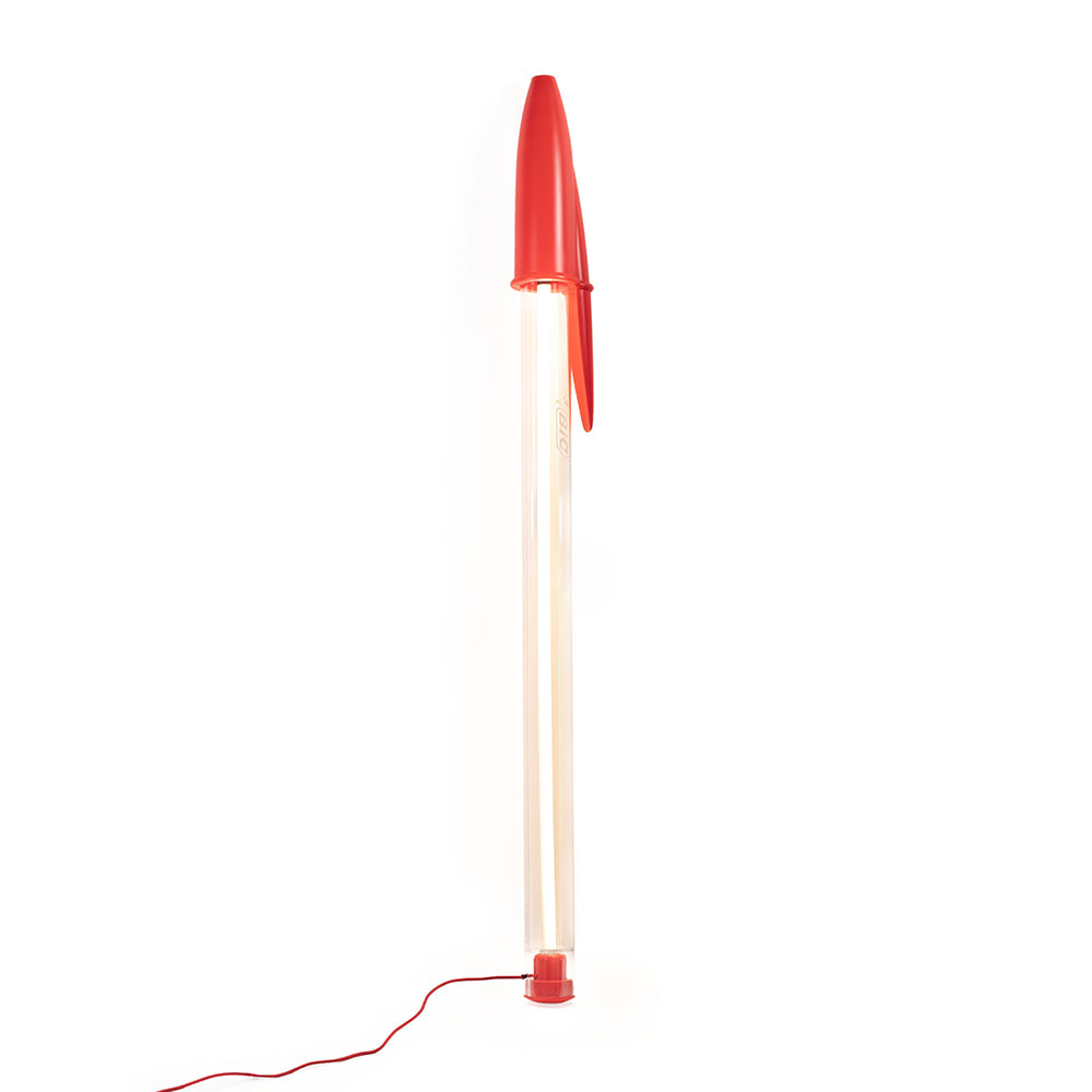 A modern floor lamp designed like a red pen, featuring a translucent clear tube body and a bright red cap and base, shown on a solid white background.