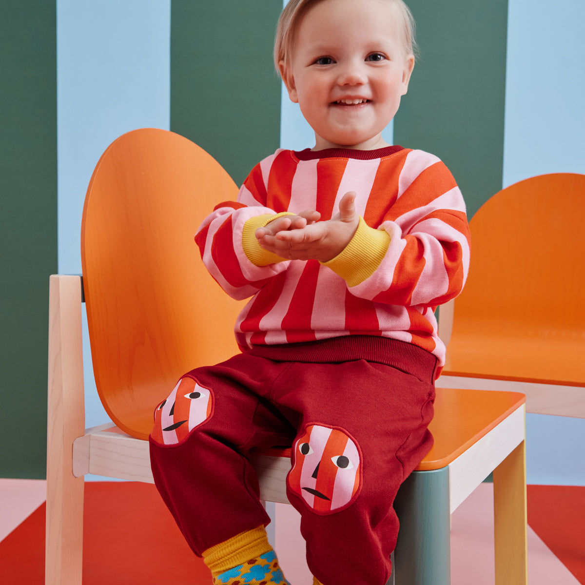 A two-piece toddler clothing set featuring a long-sleeve top with pink and red vertical stripes, yellow cuffs, and a dark red hem, paired with matching dark red pants that have yellow cuffs and a red-and-white striped circular face graphic on the lower leg.