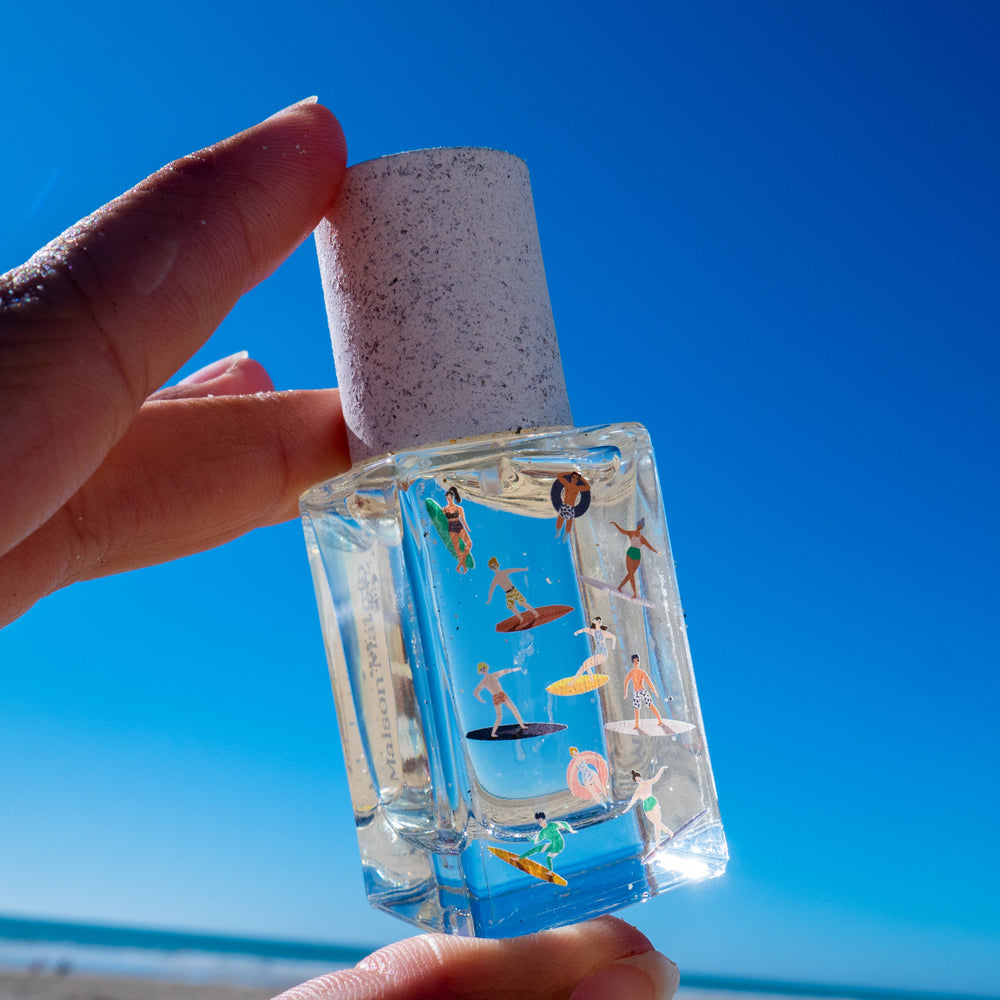 Maison Matine Bain de Midi Eau de Parfum in a clear glass bottle with colorful illustrations, displayed next to its brown kraft paper packaging box on a white background.