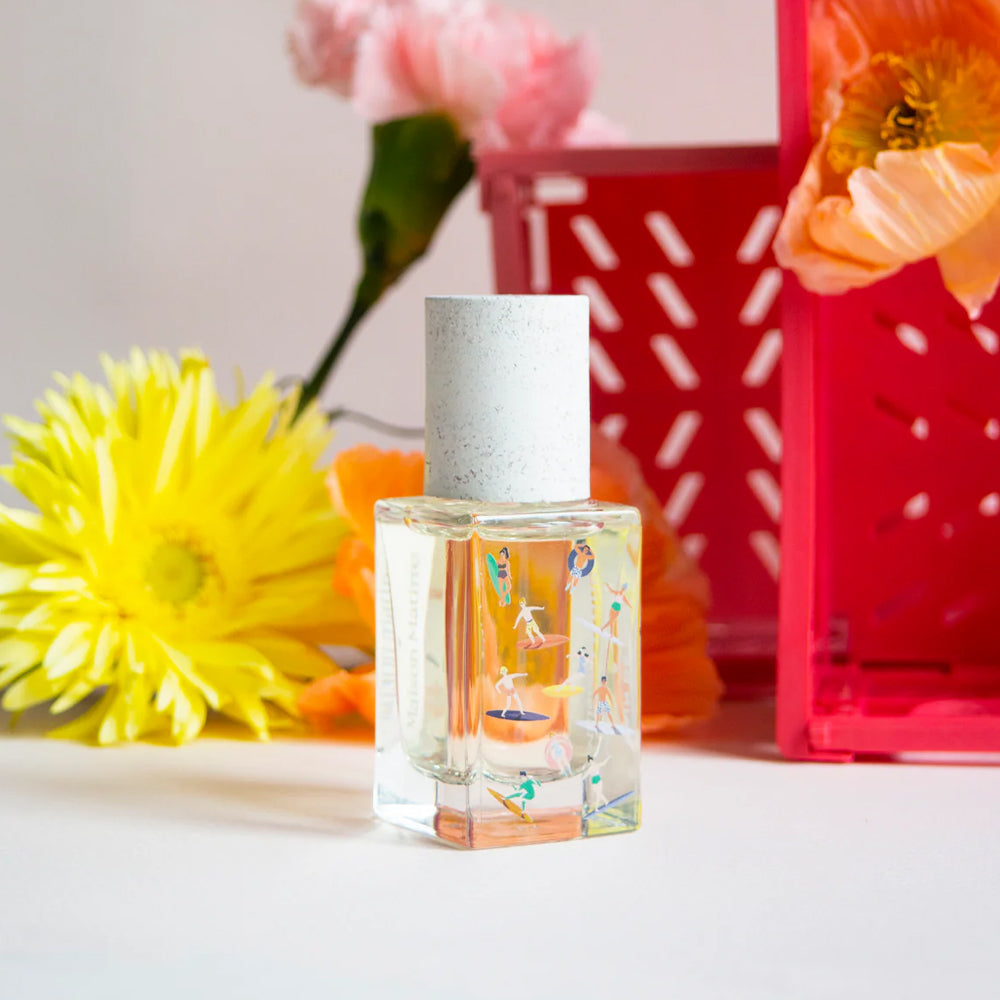 A small clear glass perfume bottle with a white cap stands on a white surface, surrounded by bright yellow and orange flowers and a pink decorative crate in the background.