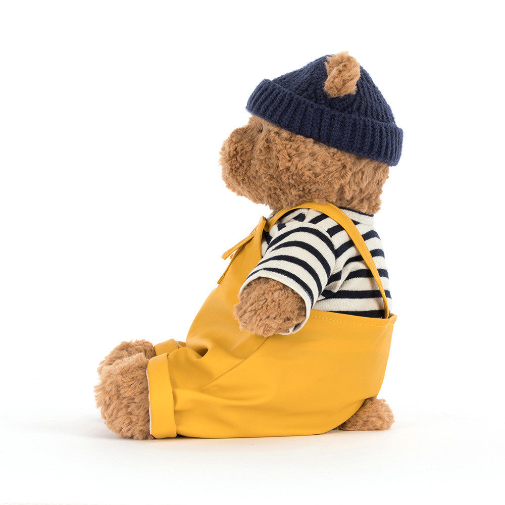 Teddy bear wearing yellow overalls, striped shirt, and navy hat on a white background