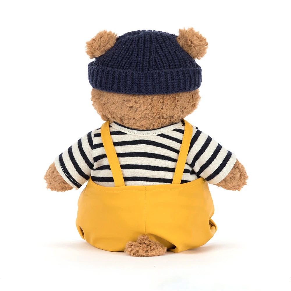 Rear view; teddy bear wearing a navy hat, striped shirt, and yellow overalls on a white background