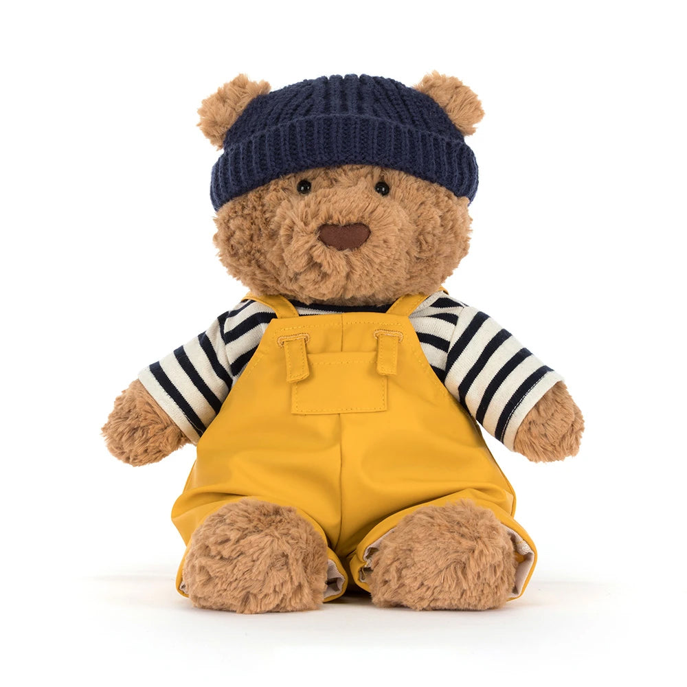 Front view; teddy bear wearing a navy hat, striped shirt, and yellow overalls on a white background