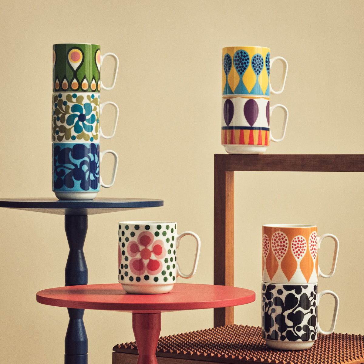 Six ceramic mugs with colorful geometric and floral patterns are arranged on small round tables against a warm beige background.