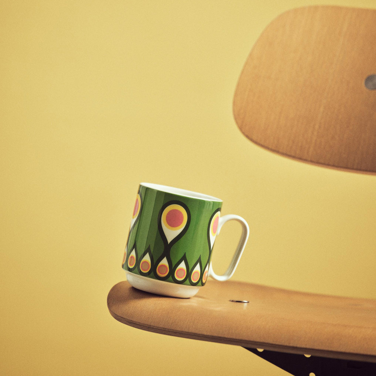A green ceramic coffee mug with a white handle and base, decorated with a pattern of orange and yellow teardrop-like shapes, resting on a wooden table next to a wooden chair against a yellow background.
