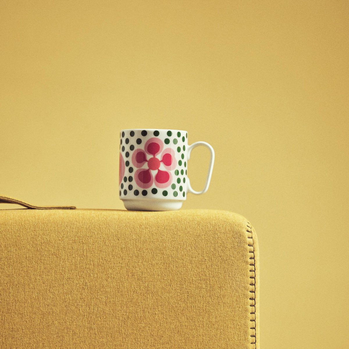A white ceramic mug with a pink floral pattern and green/black polka dots sits on the corner of a mustard yellow upholstered chair against a matching yellow background.