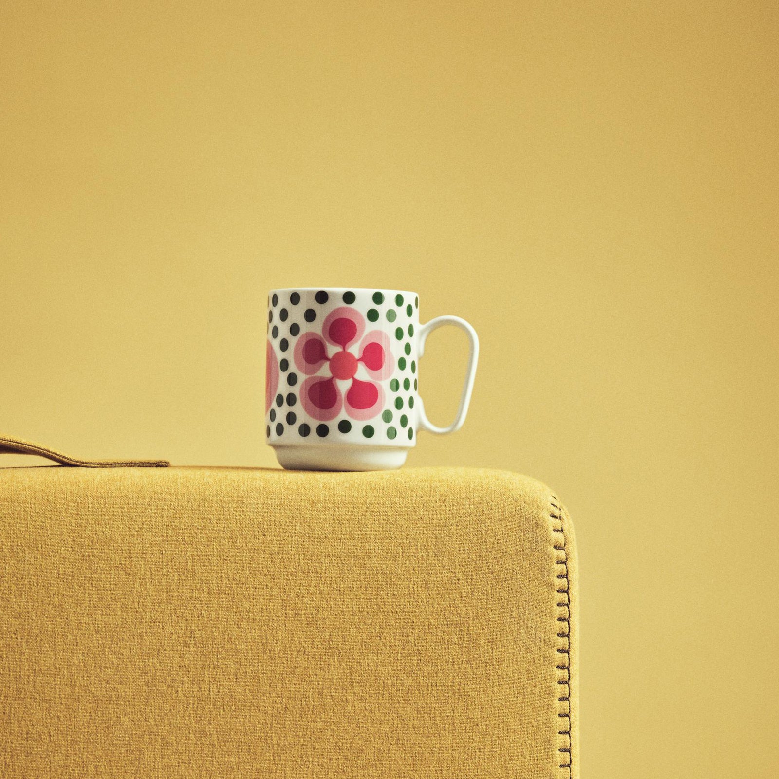 A white ceramic coffee mug featuring a retro mid-century modern design with green polka dots, a pink flower, and concentric red and orange circles, shown on a plain white background.
