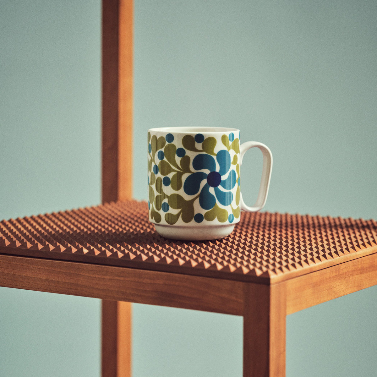 A white ceramic mug with a retro mid-century floral pattern in blue and olive green, resting on a textured wooden table against a muted blue-green background.
