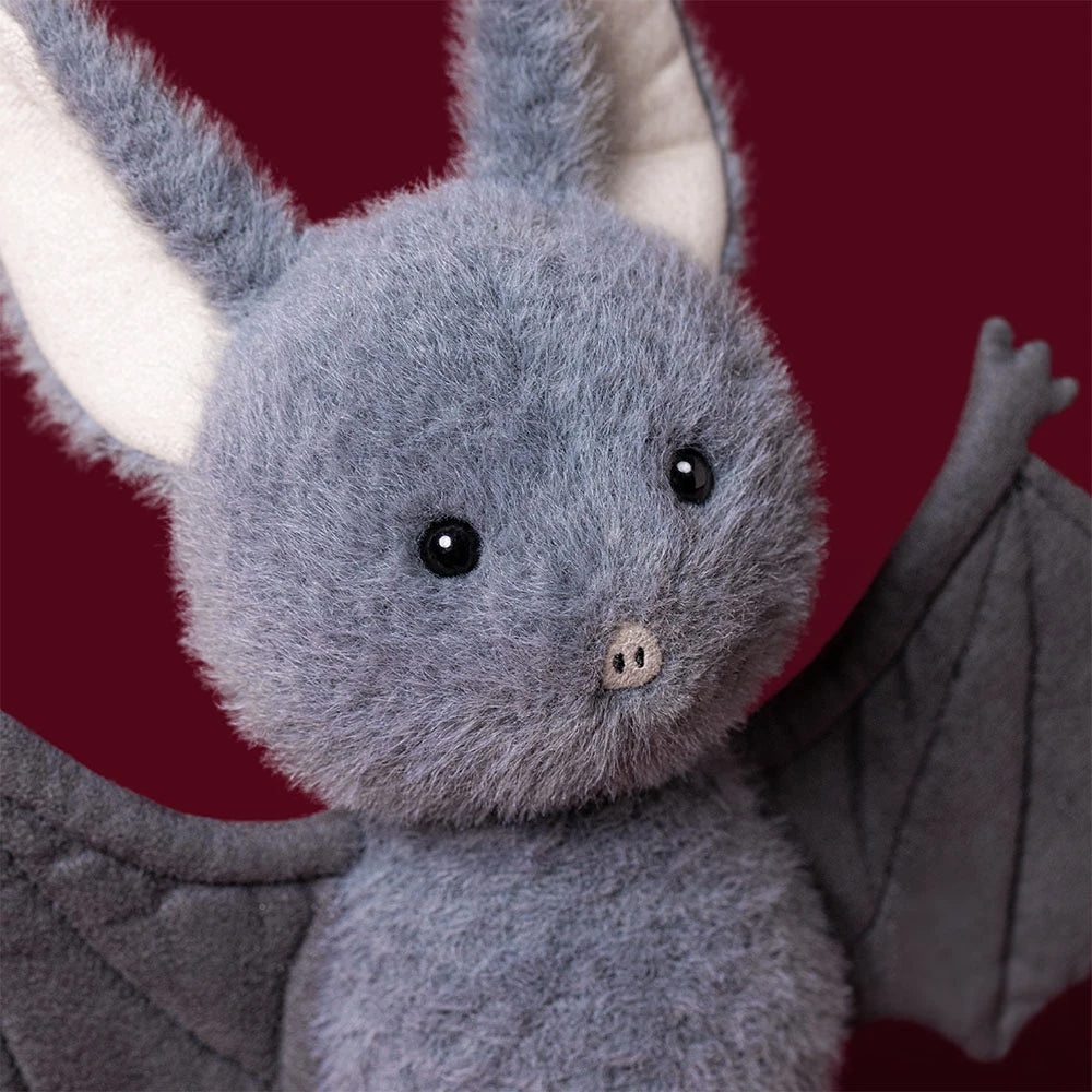 A plush bat toy with soft gray fur, large white inner ears, black bead eyes, and folded gray wings, shown against a dark red background.