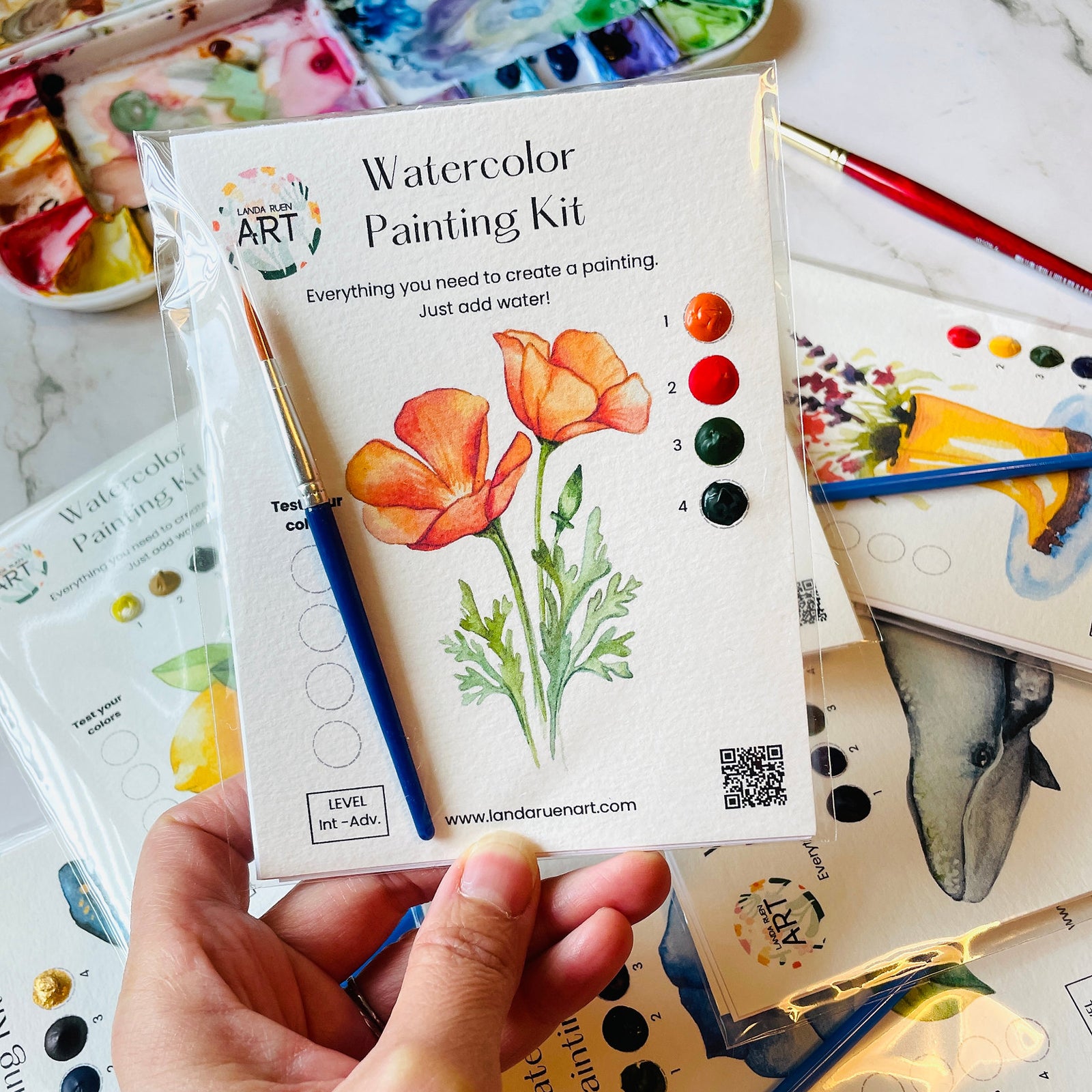A hand holds a watercolor painting kit in its clear plastic packaging, featuring orange poppy illustrations and a color wheel. The kit includes a paintbrush and is surrounded by other art supplies on a marble surface.