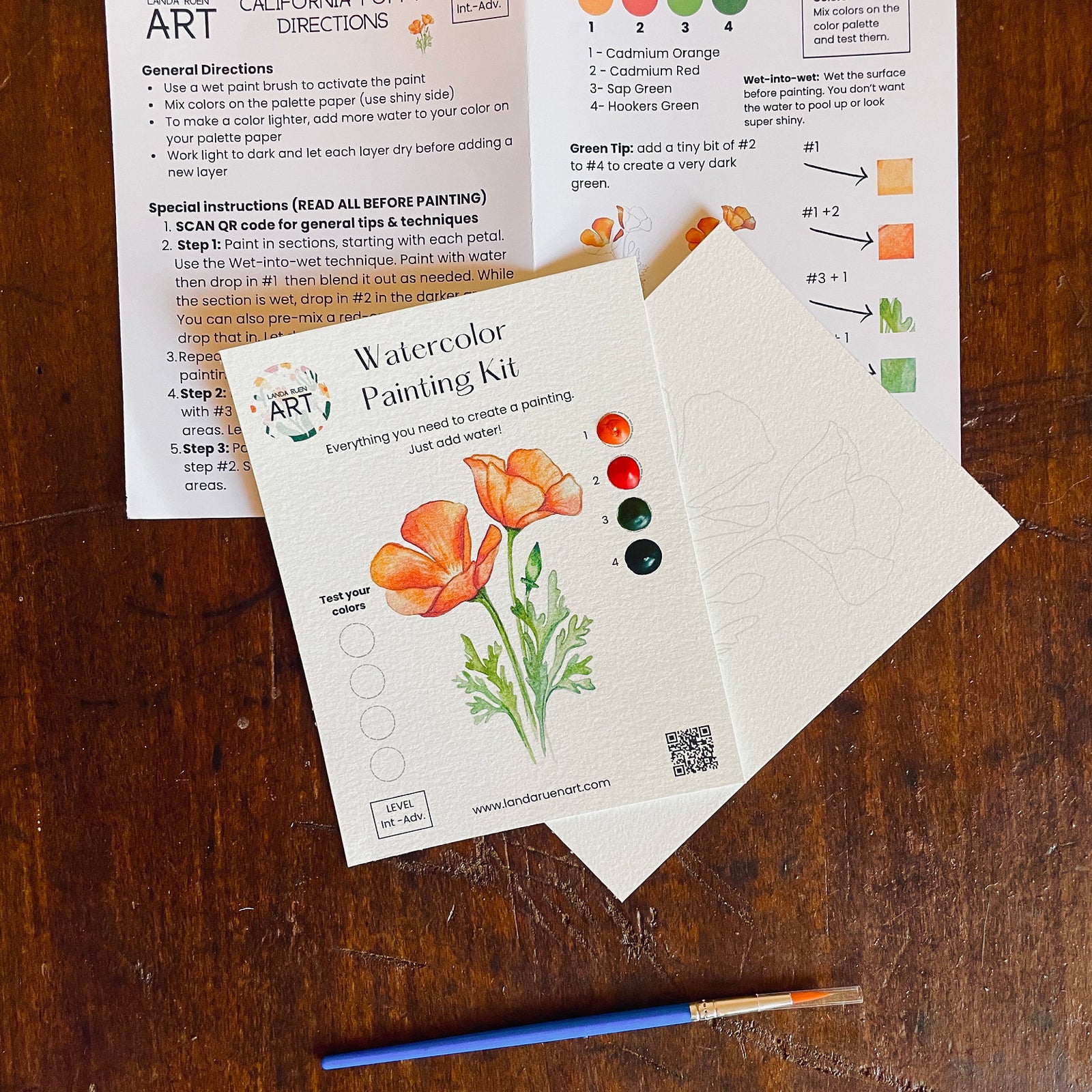 A hand holds a watercolor painting kit in its clear plastic packaging, featuring orange poppy illustrations and a color wheel. The kit includes a paintbrush and is surrounded by other art supplies on a marble surface.