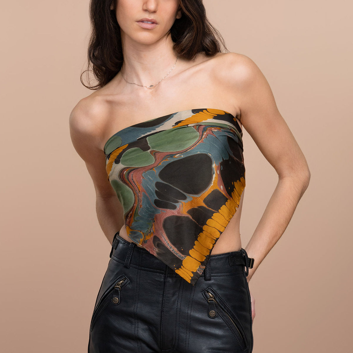A woman wearing bandana with a marbled abstract print in teal, black, olive green, and mustard yellow, paired with black leather pants against a solid beige background.