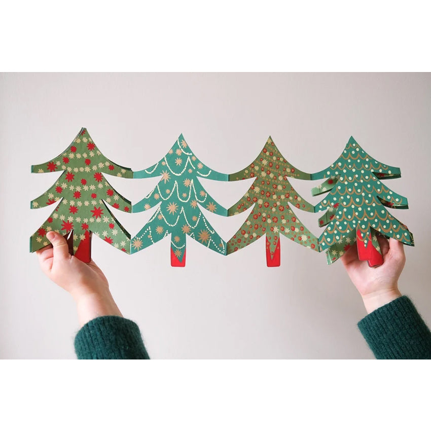 Four decorative paper Christmas tree cutouts with different holiday patterns, held up by two hands wearing green knit sleeves against a plain light background.