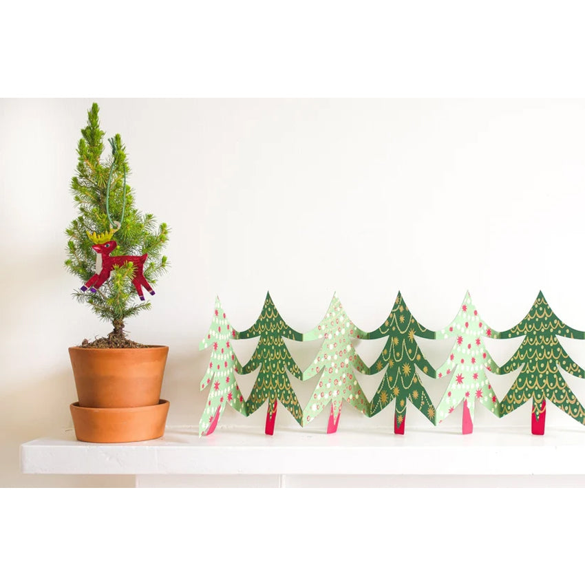 A set of six hand-painted wooden Christmas tree designs in various green shades, decorated with white dots, gold glitter patterns, and pink star accents, displayed on a white shelf next to a potted pine tree.