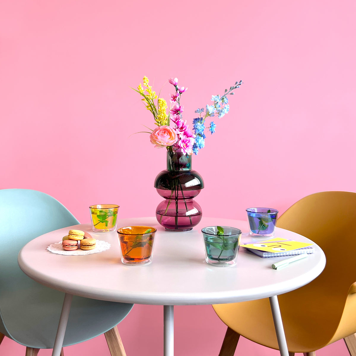 Round table with colorful glasses, a vase with flowers, and a pink background