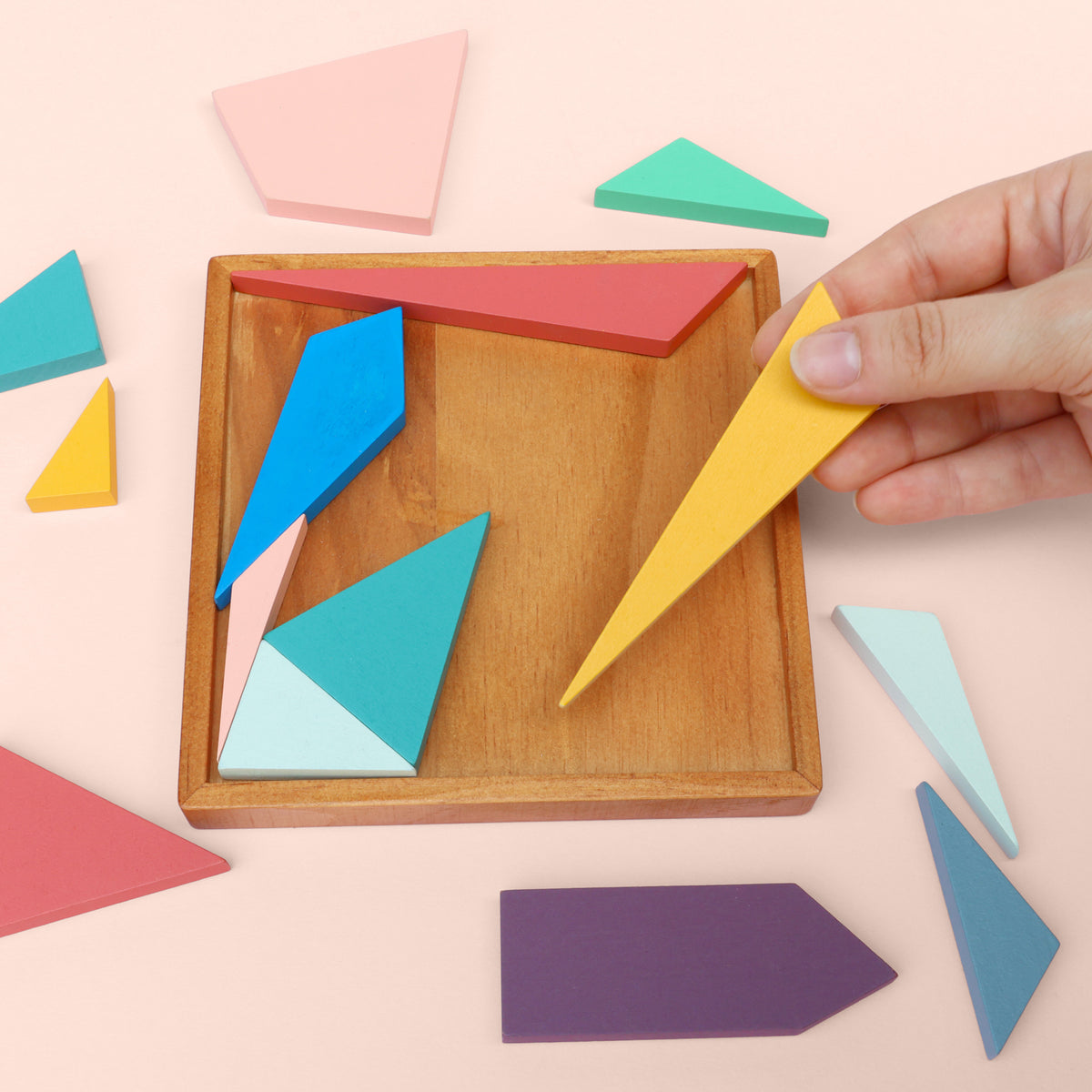 Hand placing a yellow geometric shape into a wooden puzzle box with other colorful shapes on a light pink background.