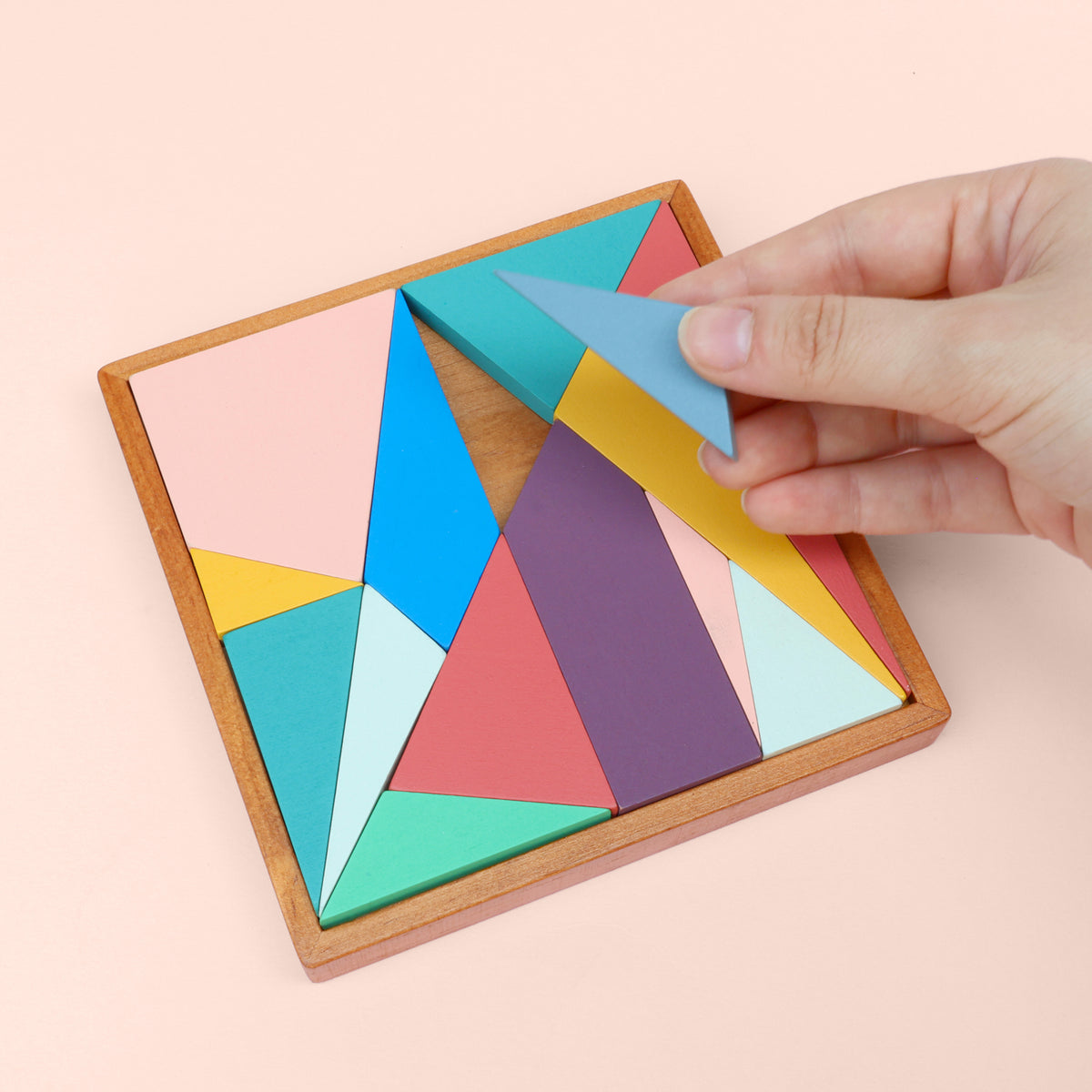 Hand placing a colorful geometric puzzle piece into a wooden frame on a pink background