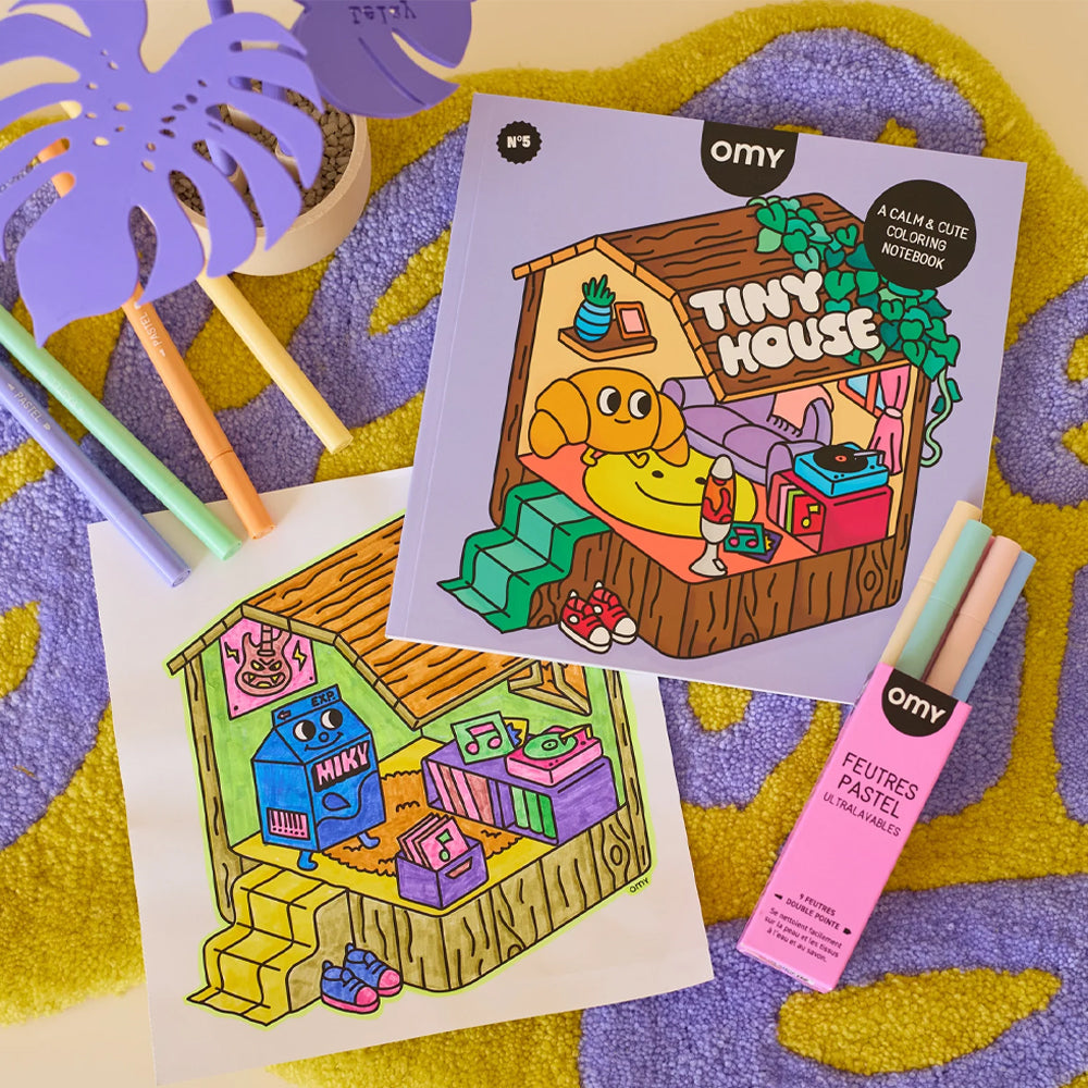 A styled flat-lay of an omy 'Tiny House' adult and cute coloring notebook, showing the open colored page and the closed cover, alongside a pink box of omy pens and pastel markers on a purple and yellow patterned rug.