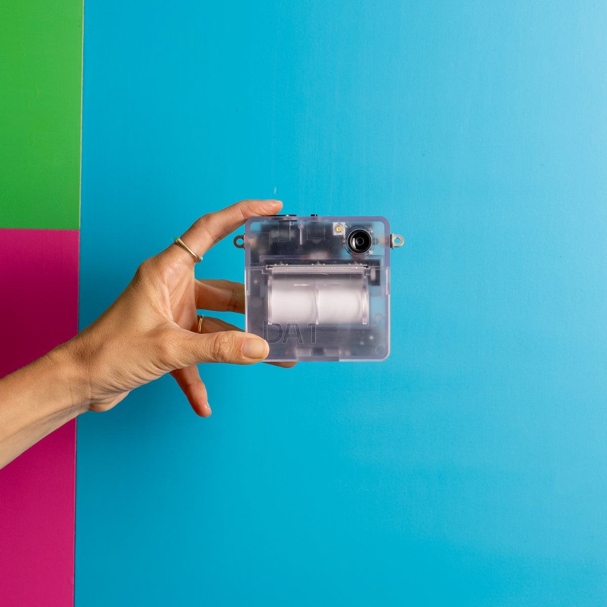 A hand holds a small transparent plastic device with a white cylindrical core against a bright blue background with a green and pink vertical stripe on the left.