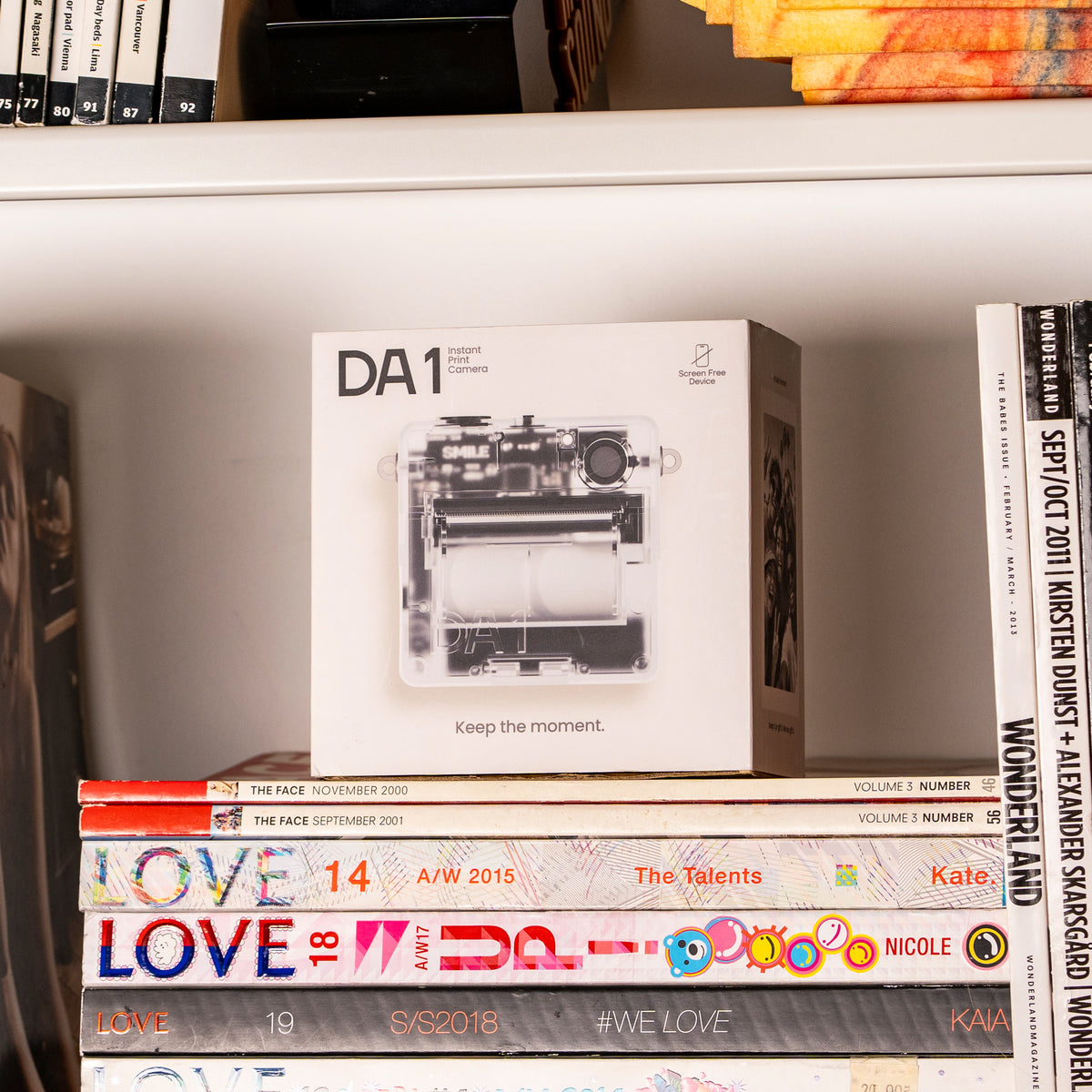 A white box for the D1 Instant Print Camera sits on top of a stack of magazines on a white bookshelf, surrounded by other books.