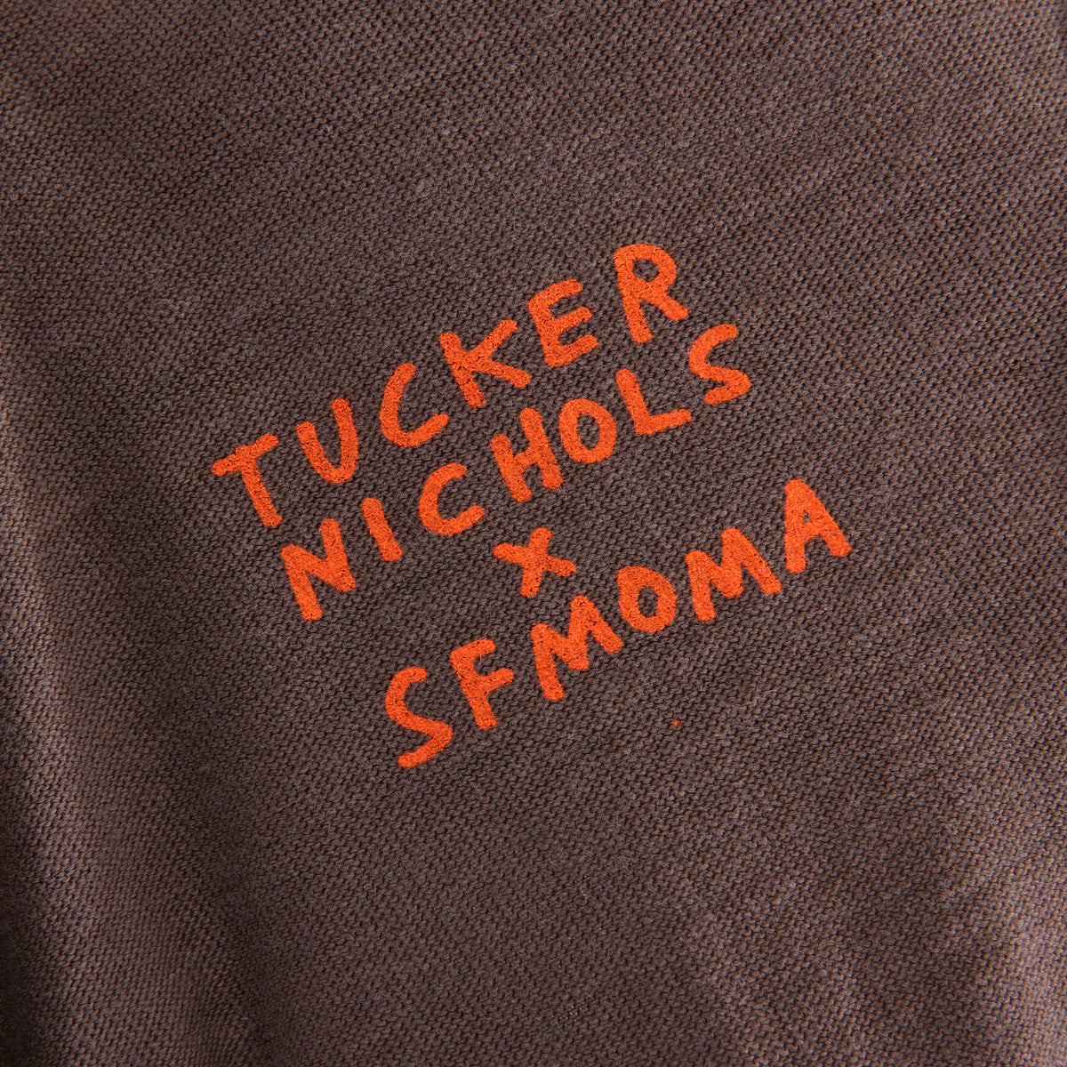 Close-up of a dark brown heathered fabric with bright orange embroidered text reading 'TUCKER NICHOLS x SFMOMA'.