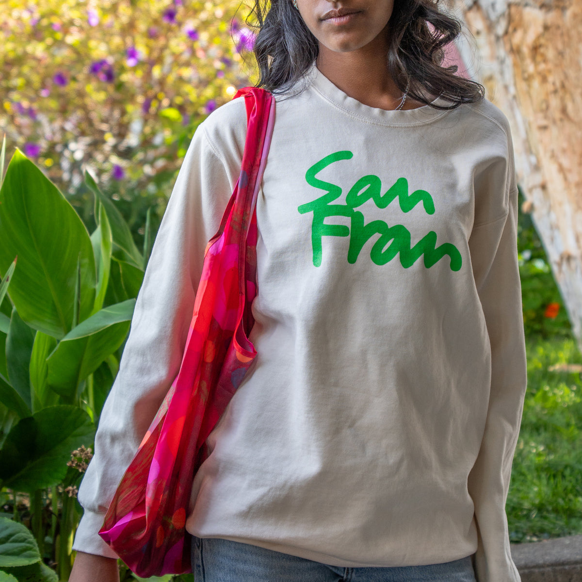 A woman wearing a cream-colored long-sleeve sweatshirt with 'San Fran' printed in green script across the chest, carrying a bright pink and red patterned scarf over her shoulder in an outdoor garden setting.