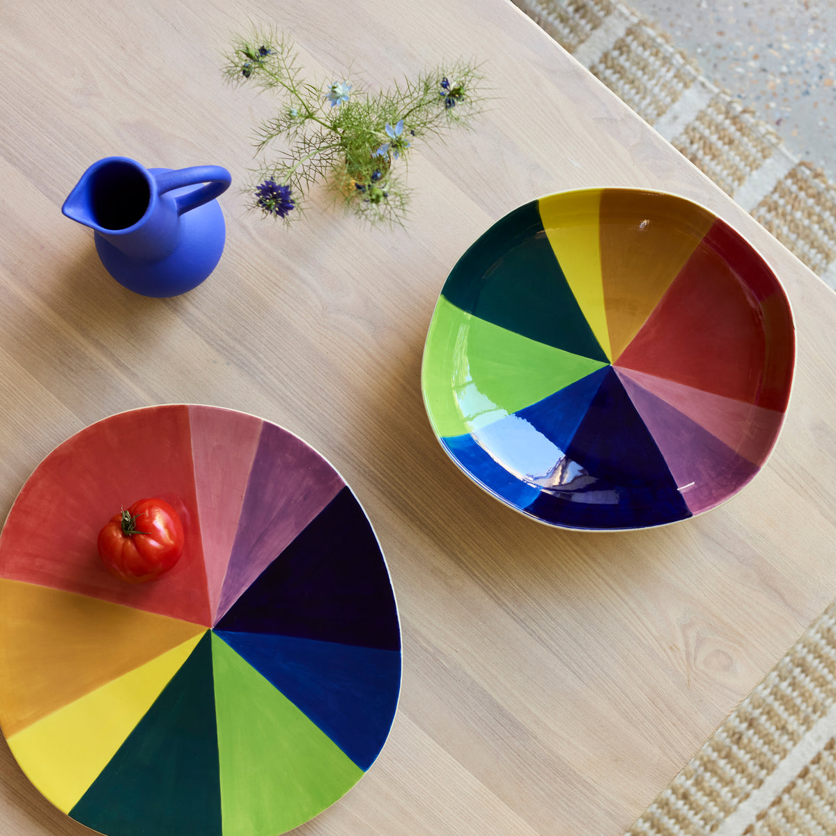 Color wheel bowl and platter with a blue pitcher and tomato on a wooden surface.