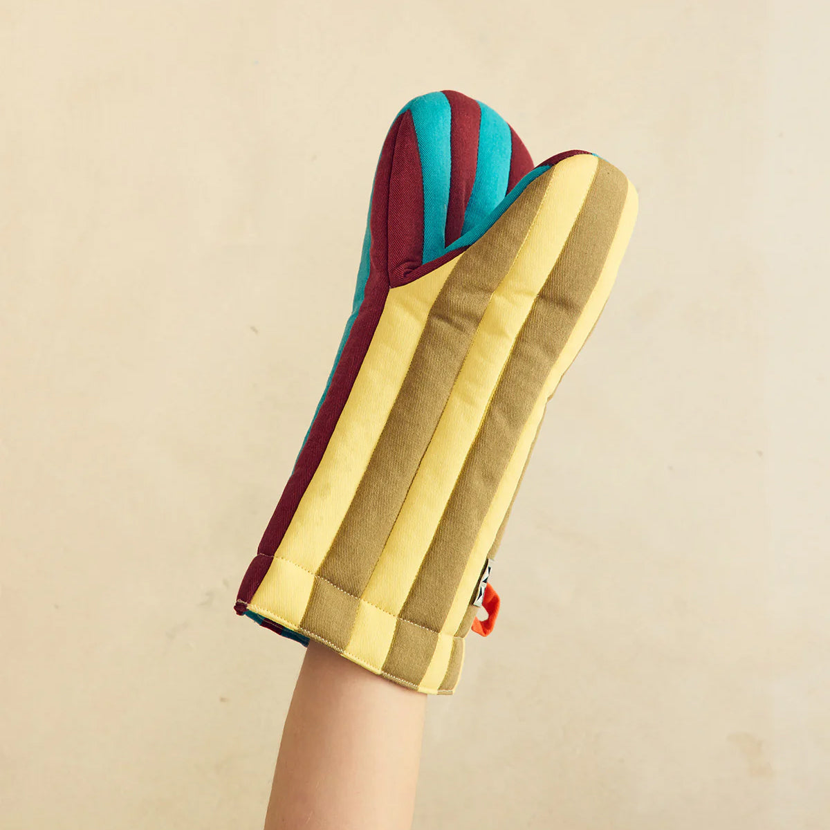 A striped fabric oven mitt held up by a person's hand against a plain beige background. The mitt features alternating vertical stripes of mustard yellow, olive green, teal blue, and burgundy red.