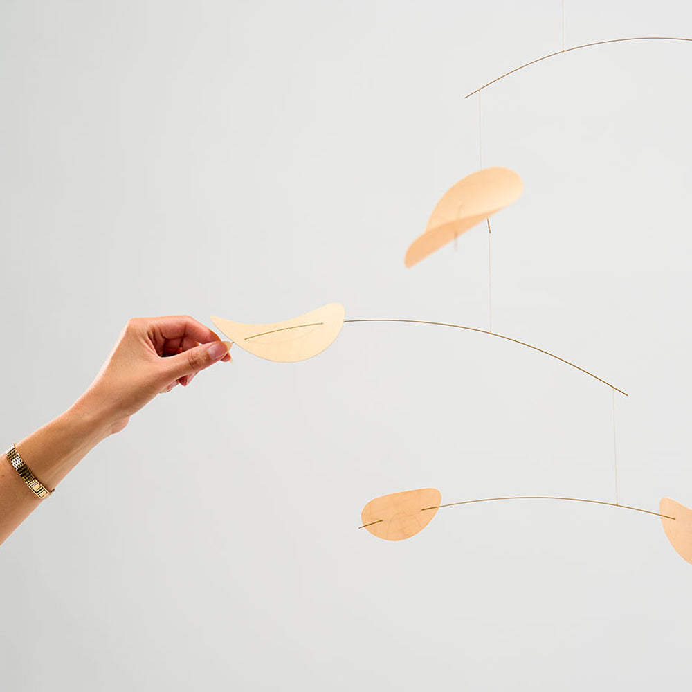 A minimalist mobile with curved gold-toned metal wires and soft peach-colored petal-like shapes, shown against a light gray background. A person's hand is holding one of the petal pieces to demonstrate scale and adjustability.