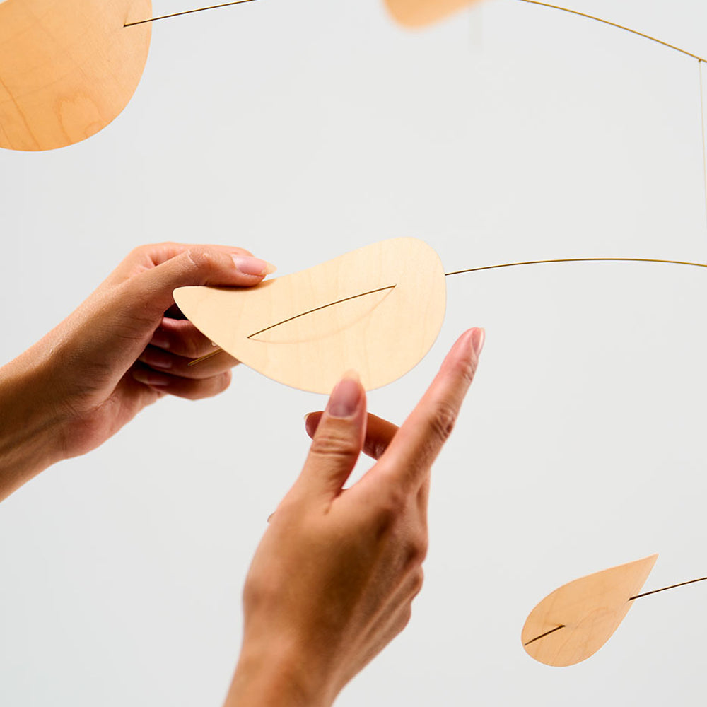 A close-up of a person's hands holding a light natural wood leaf-shaped mobile against a clean white background, with other matching wooden leaves and thin metal wires visible.