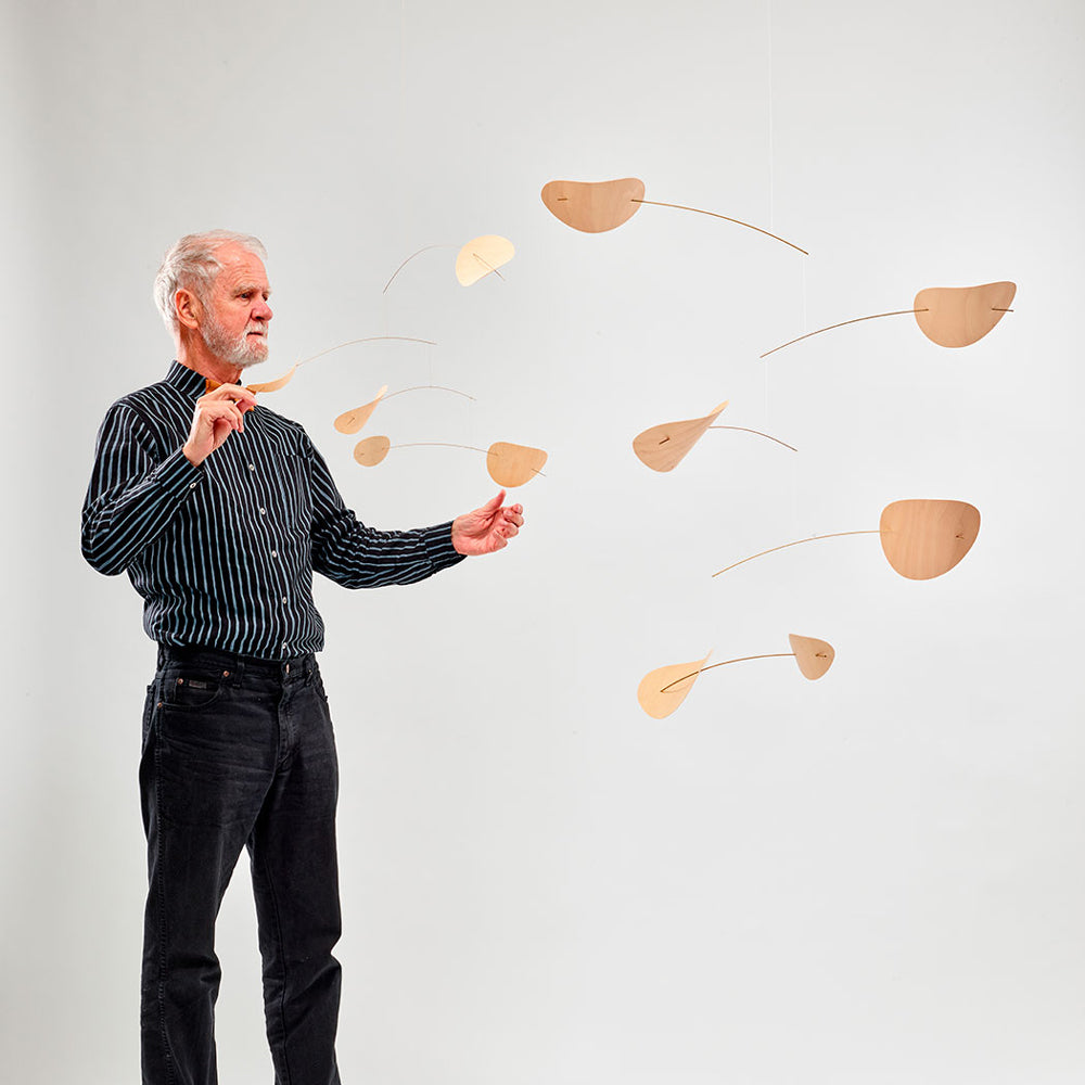 An older man with white hair and a beard stands against a light gray studio background, holding a small wooden object near several floating leaf-shaped mobiles made of natural wood. The man wears a black-and-white vertically striped button-up shirt and dark jeans.
