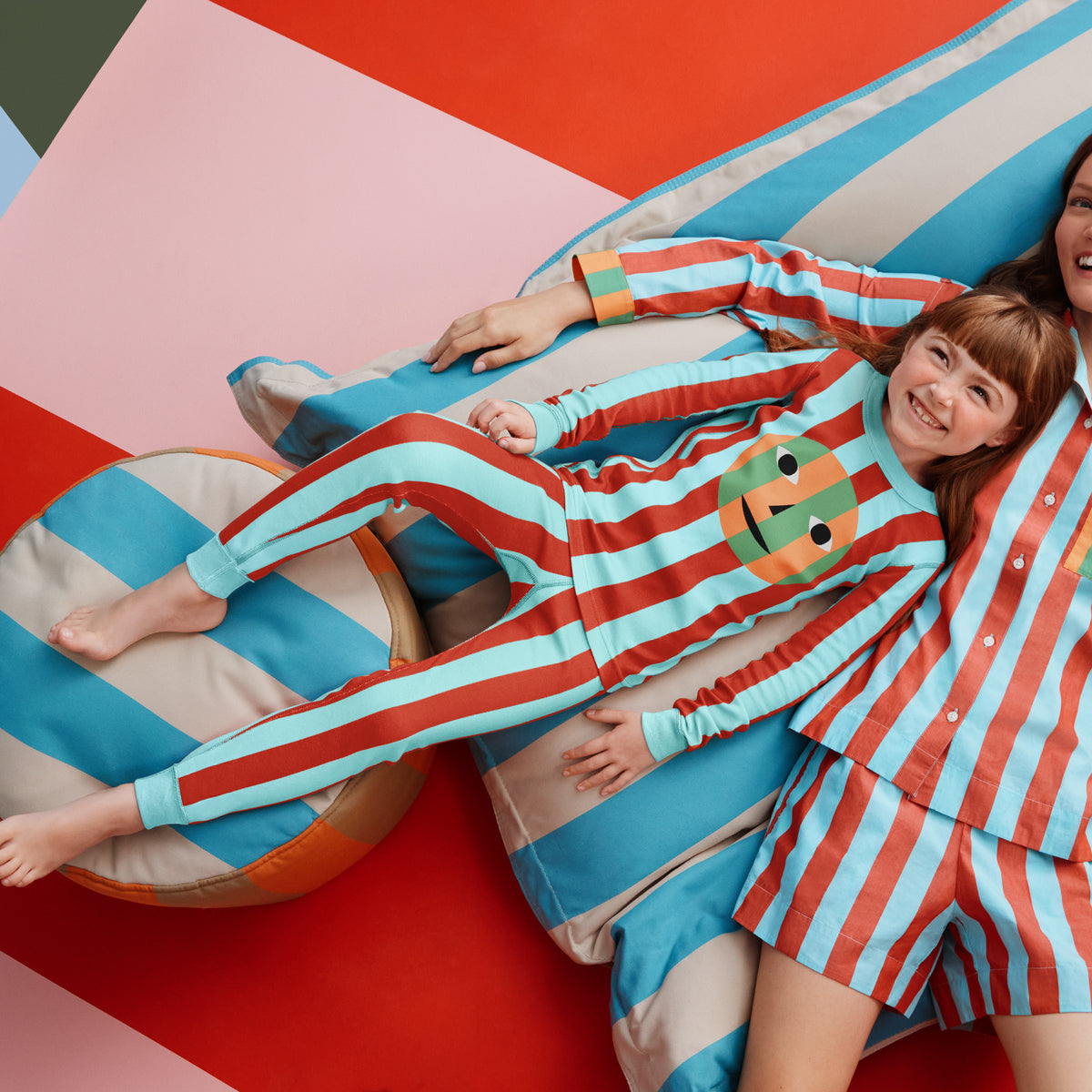 A two-piece children's pajama set featuring red and light blue vertical stripes, with a colorful abstract face graphic on the chest. The set includes a long-sleeve top and matching pants, photographed flat against a solid white background.