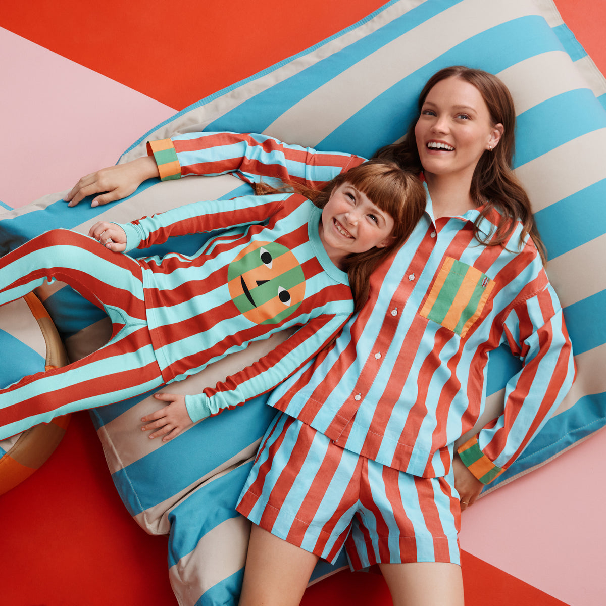 Two women lying on a striped blue, white, and beige cushion, wearing matching red and light blue striped pajamas with a colorful frog graphic on the chest.