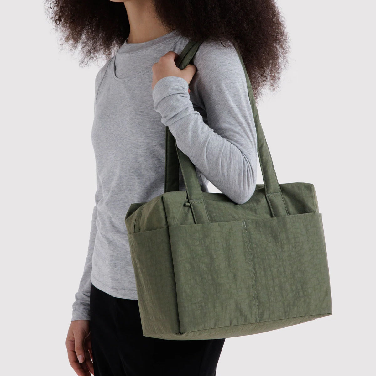 An olive green fabric tote bag with dual shoulder straps, carried over the shoulder of a person wearing a light gray long-sleeve shirt and black pants, against a plain light gray background.