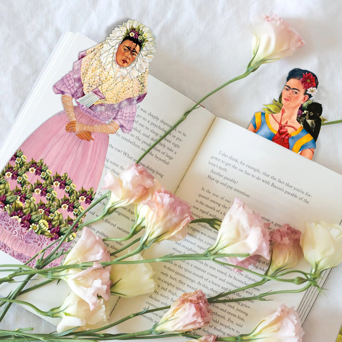 An open book on a white fabric background decorated with pale pink flowers and two illustrated paper cutouts of Frida Kahlo.