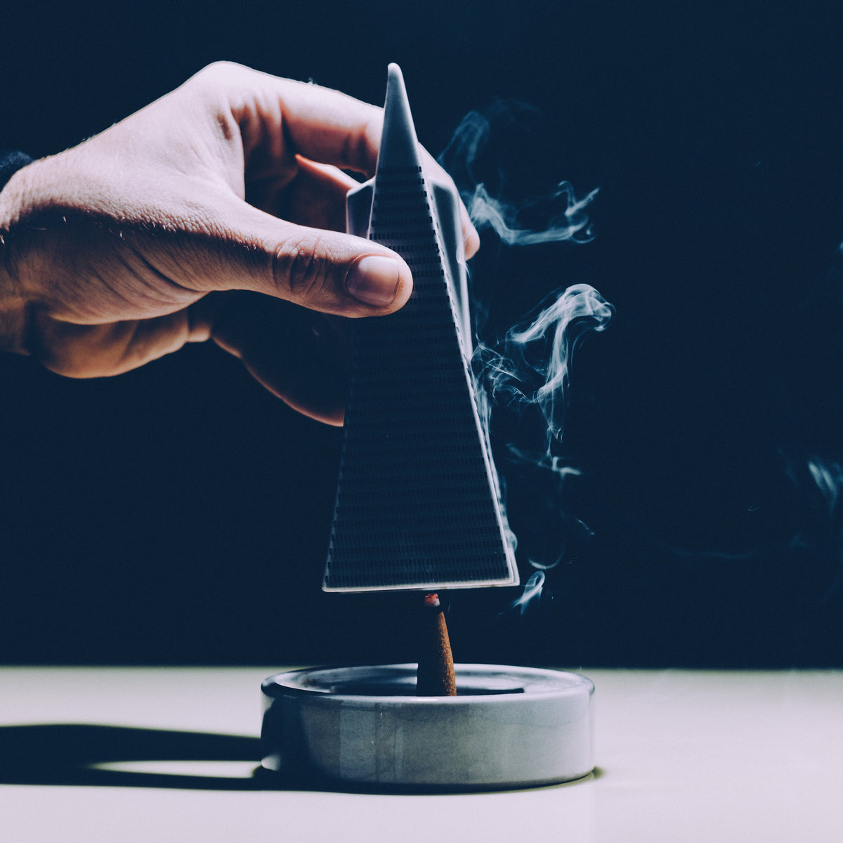 A hand holds a triangular incense holder above a small metal tray with a burning incense stick, with visible smoke against a dark background.