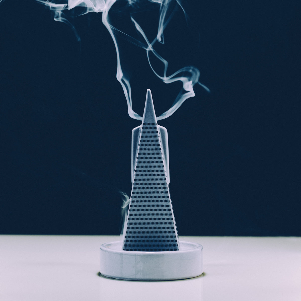 A smoking incense holder shaped like a tiered temple, with white smoke rising against a dark navy background and a white surface below.